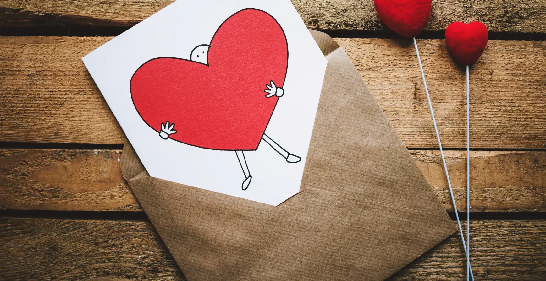 Auf diesem Bild ist ein brauner, offener Briefumschlag zu sehen. In ihm steckt eine weiße Grußkarte mit einem Strichmännchen das ein großes, rotes Herz vor sich hält.