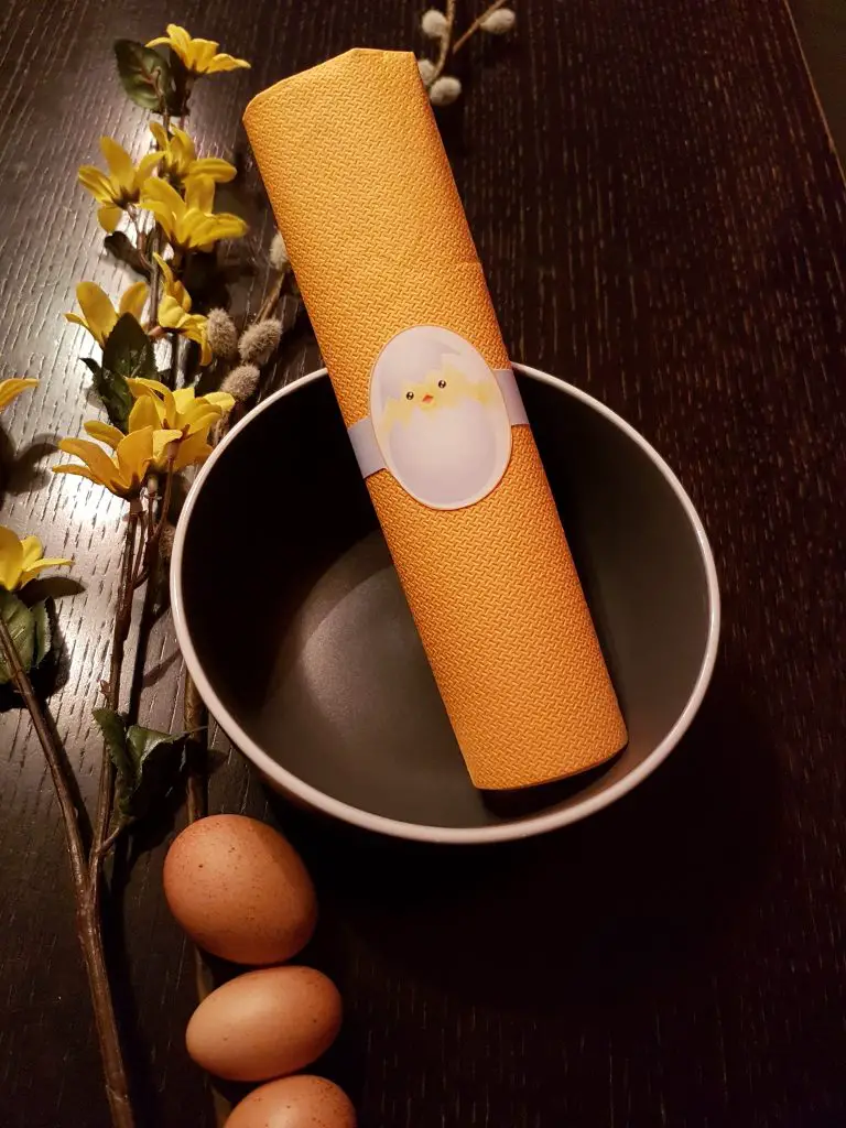 Auf diesem Bild sieht man eine orangene Serviette. Um die Serviette ist ein Serviettenring aus Papier gelegt. Auf dem Papierring befindet sich das Bild eines gezeichneten Kükens in einer lila Eischale sitzend.