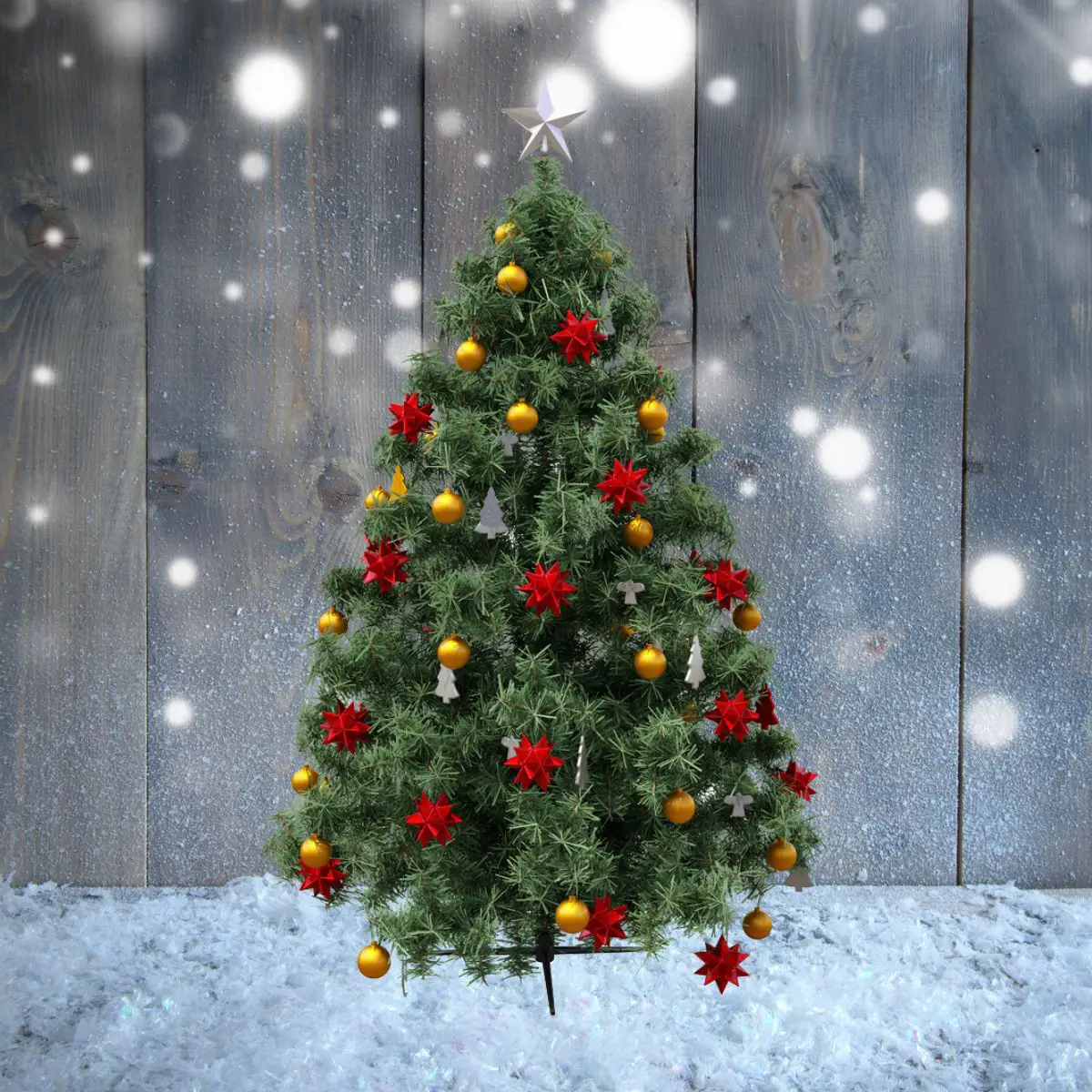 Mit roten Sternen, goldenen Kugel und weißen Tannenbäumen geschmückter Weihnachtsbaum im Schnee stehend vor einer grauen Holzwand.
